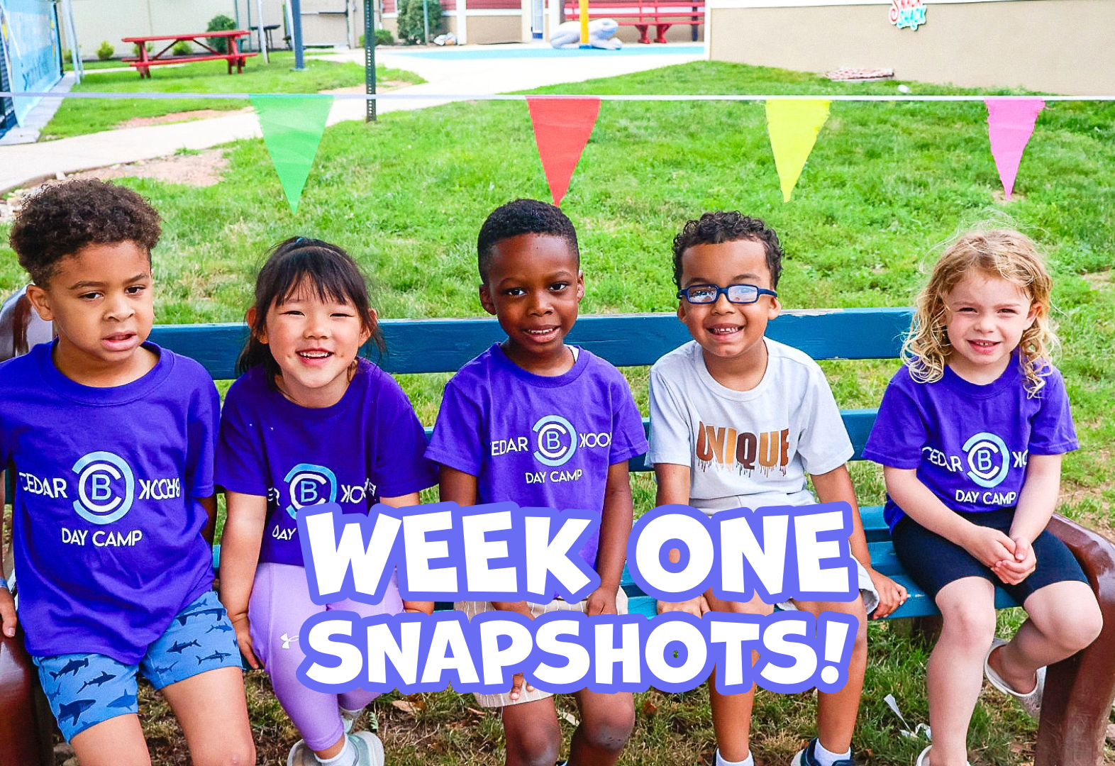 Five children sit on a bench outdoors with colorful flags and day camp shirts.