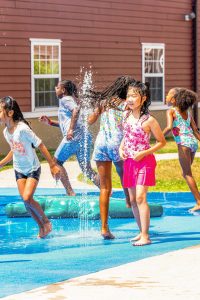 Children playing and running through water jets on a splash pad in front of a building.