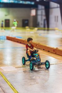 Boy in red shirt rides pedal go-kart on indoor track with orange barriers and blurred background.