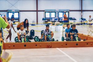 Children seated on pedal go-karts behind a starting barrier in an indoor racing area with adults nearby.