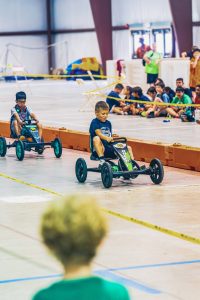 Two boys race pedal go-karts on an indoor track with children watching behind tape.