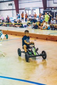 Boy in dark blue shirt rides pedal go-kart labeled RACER indoors with spectators watching.