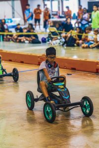 Boy with glasses and headphones driving a pedal go-kart indoors with spectators behind a barrier.