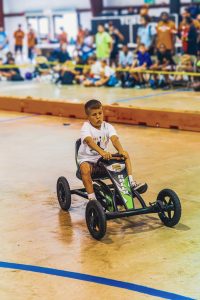 Boy in white t-shirt riding a black and green go-kart indoors on a gymnasium floor with spectators.