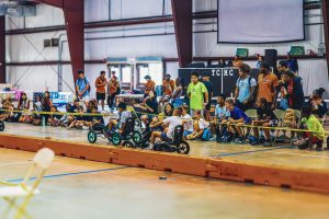 Children and teens watch and ride pedal vehicles inside a gymnasium during an event.