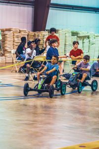 Two boys race pedal go-karts on an indoor court while others watch seated behind a barrier.