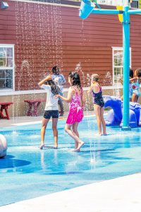 Children in swimsuits playing and splashing in a water play area with fountains and a red building.