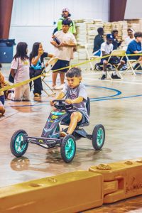 Boy riding pedal go-kart on indoor gym floor with spectators behind yellow barriers.
