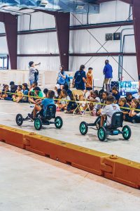 Children race pedal go-karts on an indoor track while others watch behind caution tape.