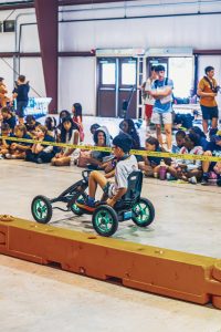 Boy riding pedal go-kart indoors with children sitting behind caution tape watching.