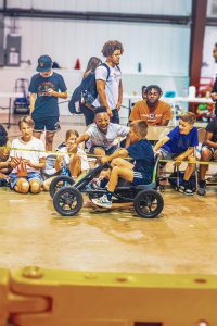 Children and teenagers play indoors with a pedal go-kart, supervised by two adults.