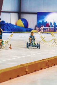 Boy riding pedal go-kart in an indoor space with chairs, caution tape, and large ball.