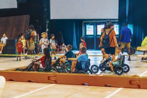 Children ride pedal go-karts inside a marked indoor track while others and staff stand nearby.