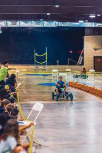 Child driving a pedal go-kart indoors on a marked track with seated spectators nearby.