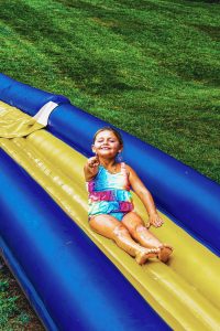 Girl in a colorful swimsuit sliding down a yellow and blue inflatable water slide on grass.