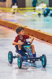 Boy in red shirt riding a pedal go-kart indoors on a smooth surface near barriers.