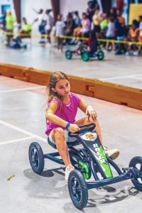 Girl in pink shirt riding pedal go-kart labeled Racer on indoor track with spectators behind barrier
