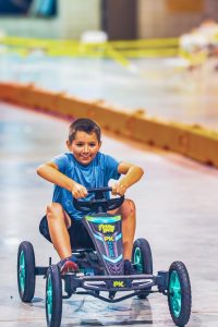 Boy in blue shirt riding pedal go-kart on smooth indoor track with safety barriers.