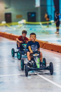 Two boys riding pedal go-karts on an indoor track with orange barriers and people in background