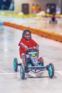 Child in red shirt riding a black and green pedal go-kart indoors on a concrete floor.