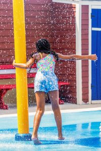 Girl in floral top and blue shorts playing under water spray from yellow pole at splash pad.