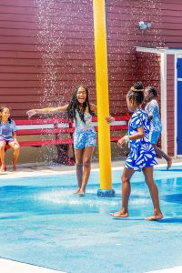 Children playing in water sprays at an outdoor splash pad near a red building with a blue door.