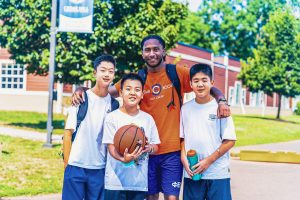 Man in orange shirt with backpack standing with three boys holding basketball and water bottle outdoors.