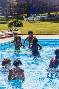Boys playing and splashing in a sunny outdoor swimming pool with lawn chairs and umbrellas nearby.