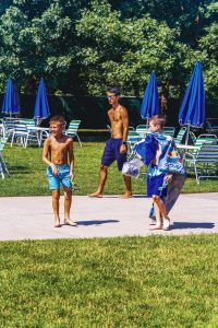 Three boys in swimwear walk on a path beside grass and outdoor chairs with blue umbrellas.