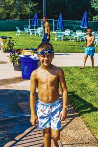 Boy wearing swim trunks and goggles on forehead standing near poolside on a sunny day.