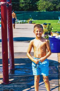 Boy in blue swim trunks holding goggles standing near wet concrete and red poles at poolside.