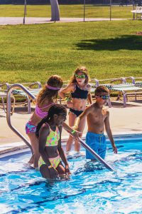 Four children in swimwear and goggles entering a sunny outdoor swimming pool near lounge chairs.