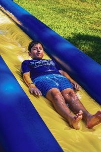 Boy in blue outfit sliding down a yellow and blue inflatable water slide on grass.
