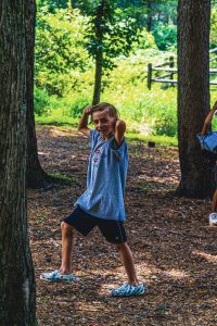 Boy in gray shirt and black shorts posing playfully in a sunny wooded area.