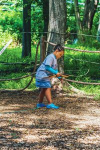 Child in gray shirt, blue skirt, and shoes holding a bow and arrow in a wooded archery area.