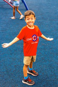 Boy in red Cedar School Day Camp t-shirt and khaki shorts standing on blue playground surface with arms extended