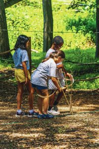 Three children outdoors on wood chip ground with a bow and arrow in a wooded area.