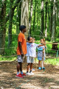 Man in orange shirt teaches two children archery in a wooded, roped-off area with wood chip ground.