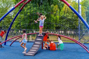 Children playing on a colorful playground climbing structure with blue rubber flooring and trees nearby