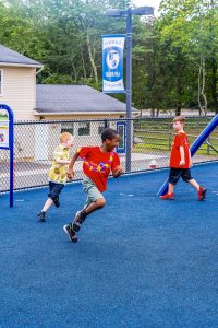Children playing on blue playground surface near fence and Cedar Hill Preparatory School banner.