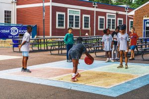 Children playing with a red ball on a colorful court outside red and brick buildings at a day camp.