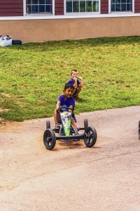 Two boys on grass near a building, one riding a pedal go-kart on a paved path