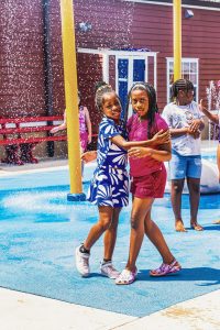Children playing on a blue surface with water sprays and yellow poles near a red building.