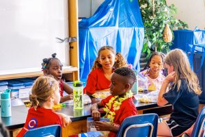 Six children sitting around a table in a classroom eating snacks and drinking from water bottles.
