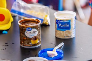 Two open containers of chocolate and vanilla frosting on a table with a spoon on a blue lid.