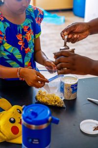 Child holding popcorn bag while another scoops chocolate spread near frosting and Pikachu hat.