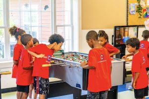 Children in red shirts playing foosball in a classroom with windows and a TV.