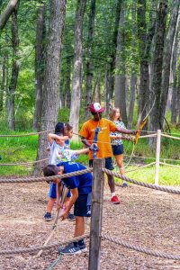 Children and instructor practicing archery in a wooded area enclosed by ropes and posts.