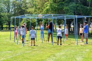 Children and adults playing a ball game around a large blue metal frame in a grassy park area.