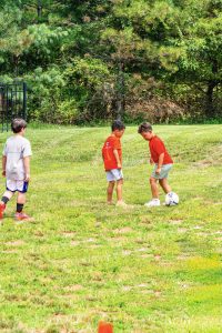 Three boys playing soccer on a grassy field near trees, two in red shirts, one in gray.